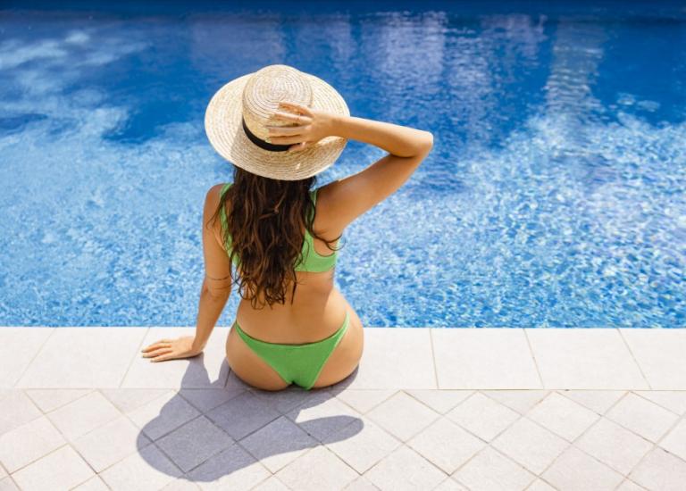 Donna in bikini verde e cappello seduta a bordo piscina.