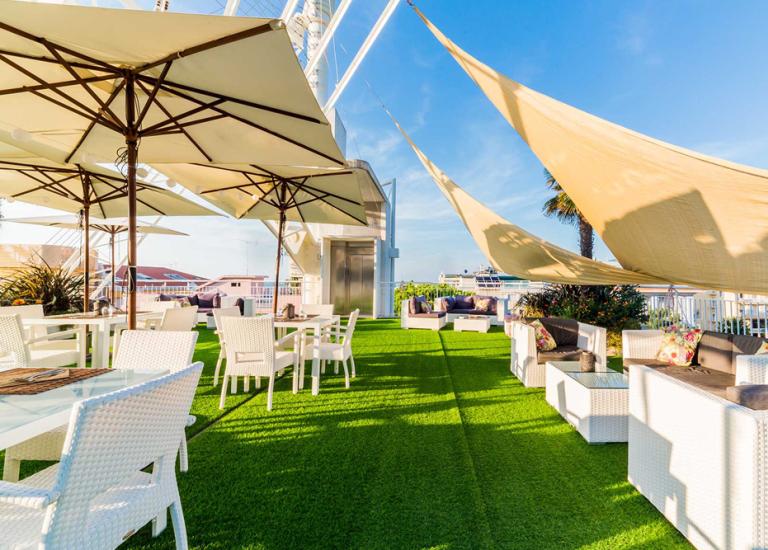 Terrazza soleggiata con tavoli, sedie bianche e ombrelloni su prato verde.